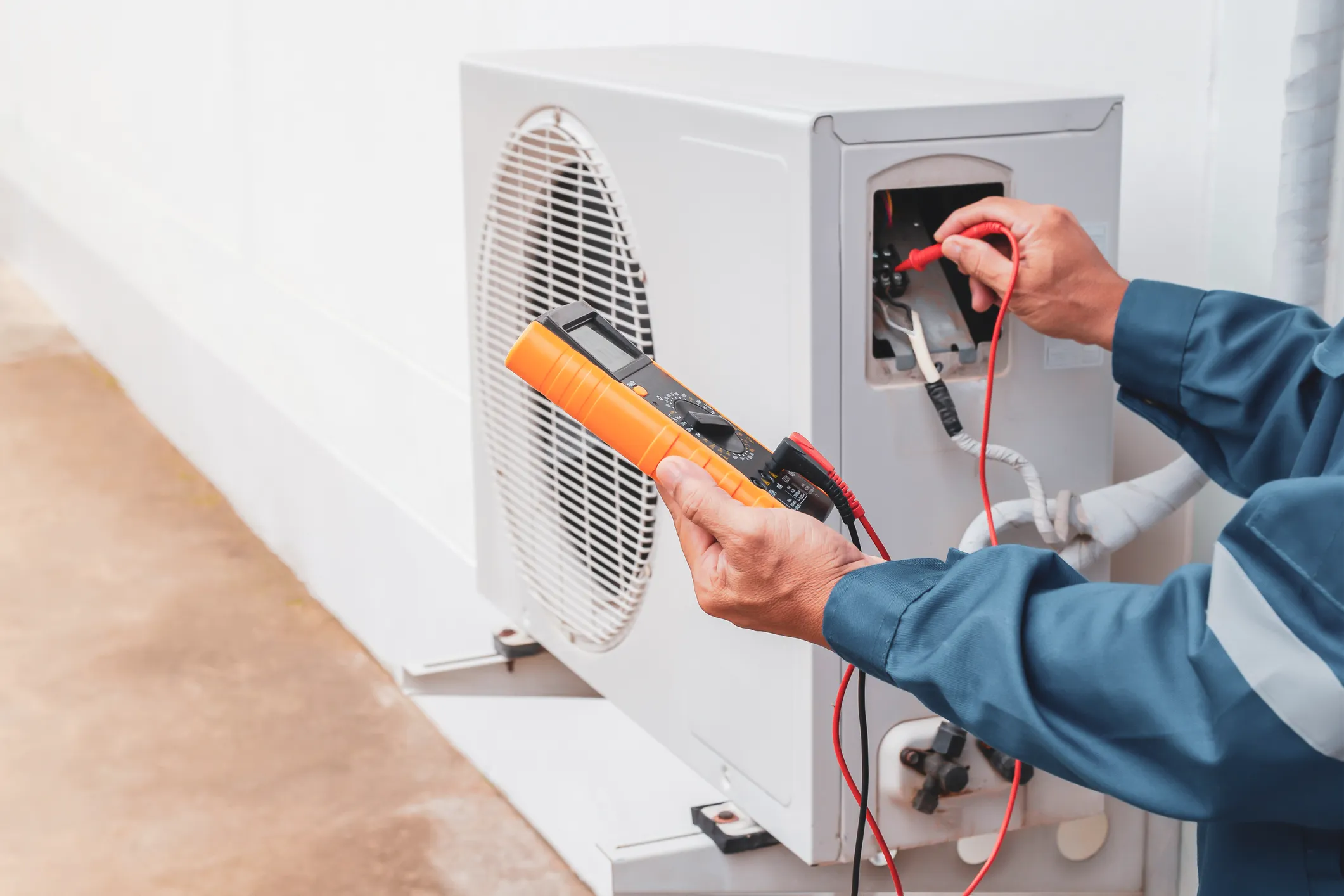 A technician in blue work clothes is using a multimeter to test the wiring of an outdoor air conditioner.