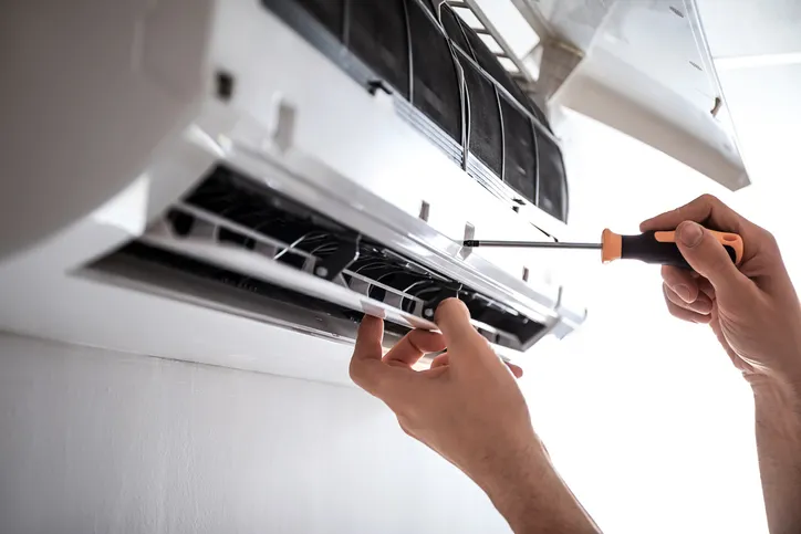 A close-up of a technician's hands using a screwdriver to perform maintenance on a wall-mounted air conditioner.