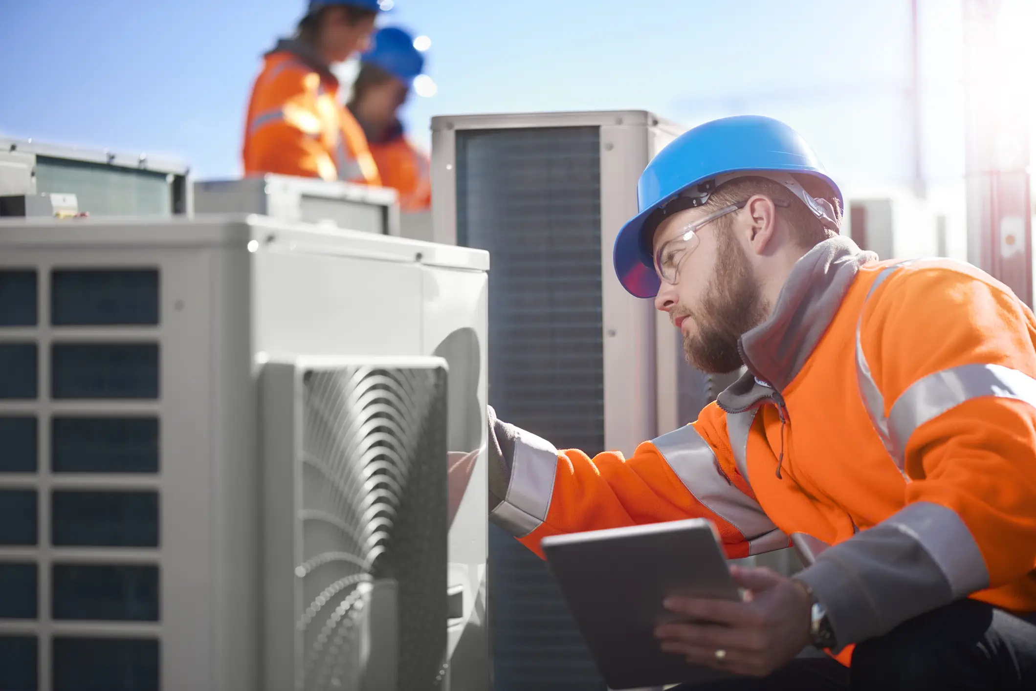 A focused male HVAC technician, wearing a bright orange jacket and a blue hard hat, inspects an outdoor air conditioning unit while holding a tablet. In the background, two other workers in similar safety gear are partially visible near more AC units on a sunny rooftop.
