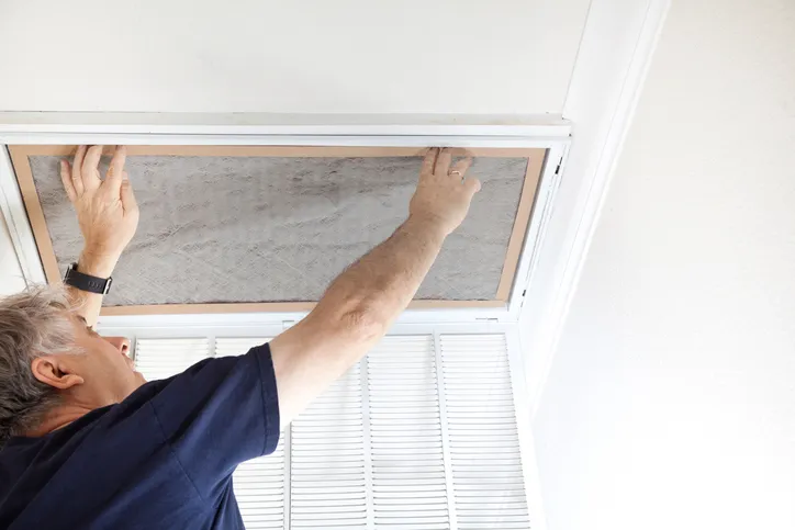 A person's hands are replacing a dusty, gray air filter in a ceiling vent.