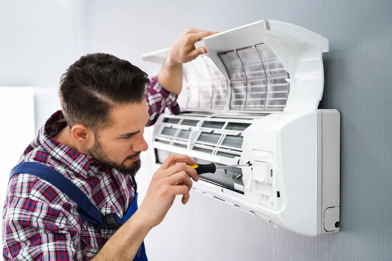 A technician with a beard and plaid shirt is using a screwdriver to fix a wall-mounted air conditioner.