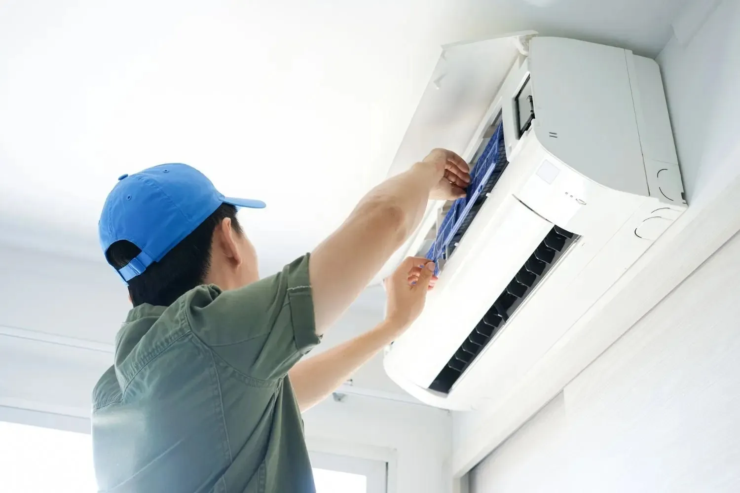 A technician wearing a baseball cap inspects the filter of a white, wall-mounted indoor AC unit.