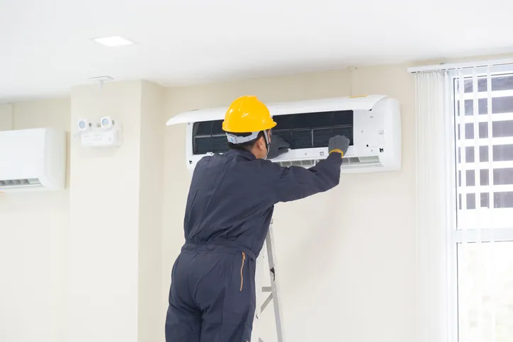 A technician in a hard hat standing on a ladder, servicing a wall-mounted air conditioner in a room.