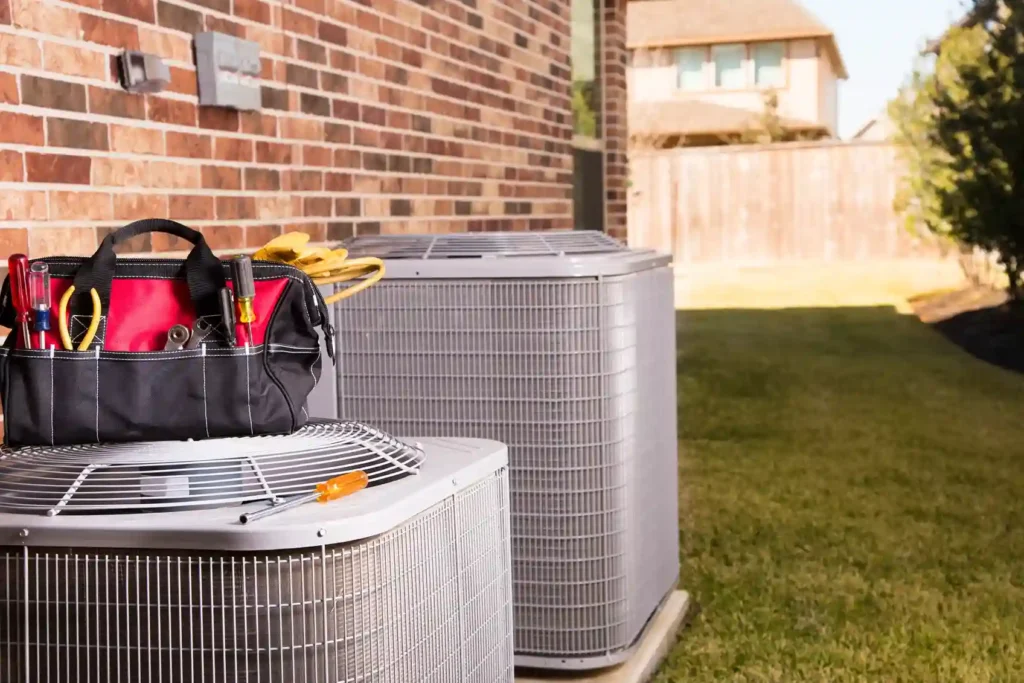 Two outdoor air conditioner units are shown with a red and black tool bag on top of the closer unit.
