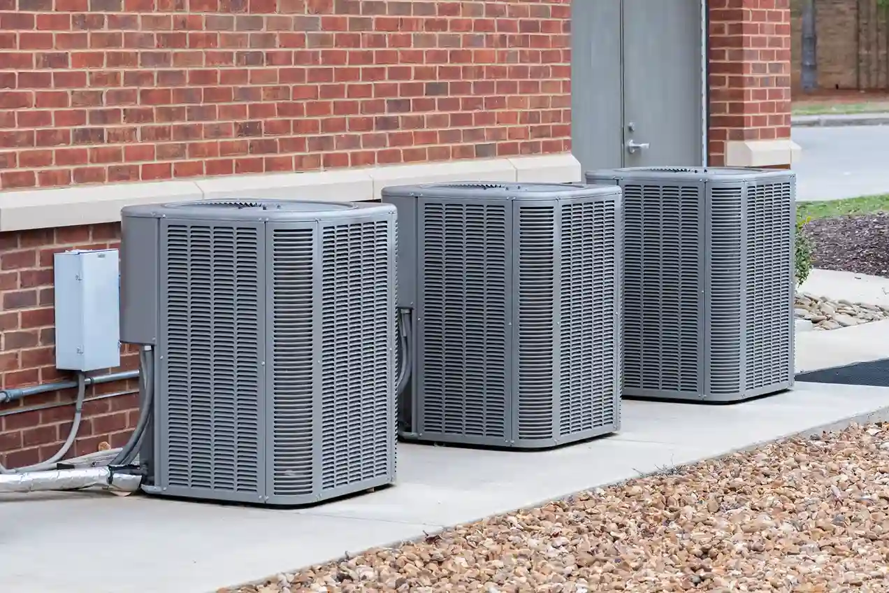 Three large, gray outdoor air conditioner units are installed in a row next to a brick building.