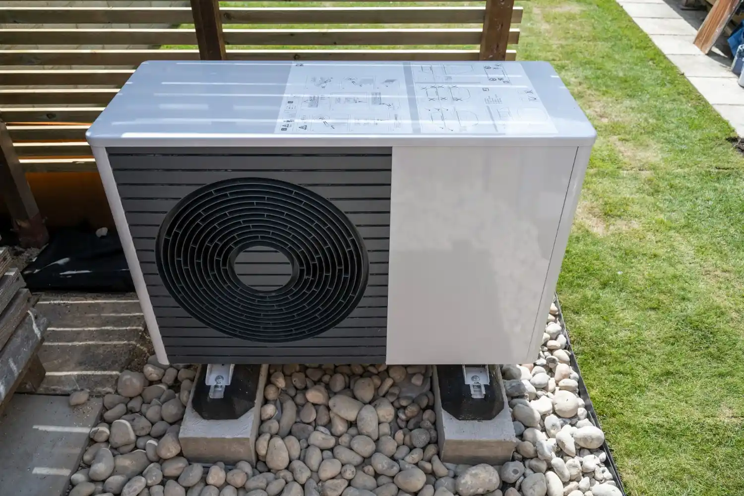An outdoor air source heat pump unit, white and gray, sitting on supports surrounded by river stones and grass.