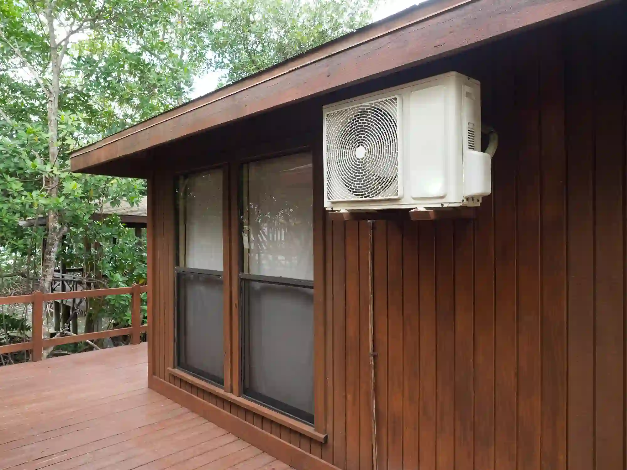 A white ductless mini-split outdoor condenser unit is mounted high on the brown wood-paneled exterior wall of a building. Below the unit are two vertical windows. The structure features an overhanging roof and a wooden deck and railing are visible below and to the left, surrounded by trees.