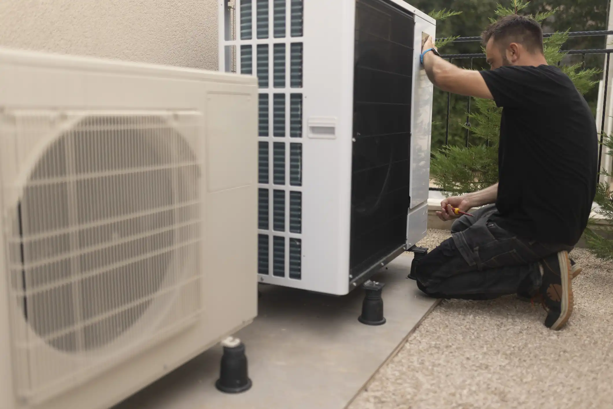 A technician in a black shirt and gray pants is kneeling and performing service on an outdoor heat pump unit, while another identical unit stands next to it. They are both elevated on black risers on a paved area next to a building.